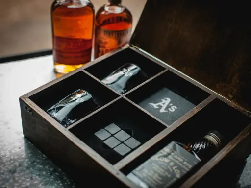 Elegancki zestaw z whisky w drewnianej skrzynce: dwie butelki, kieliszki z monogramem "A's", kamienie do whisky i podkładka.