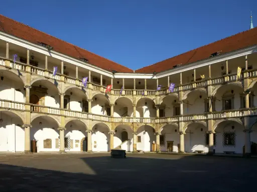 Dziedziniec renesansowego zamku z arkadowymi krużgankami, ozdobiony flagami.