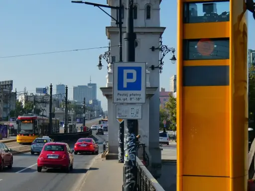Żółty fotoradar stoi przy drodze, gdzie widać ruch samochodowy i tramwaj. Obok znak parkingu płatnego.