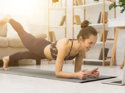 Uśmiechnięta kobieta w desce z nogą w górze, oglądająca trening na laptopie. To świetny sposób, jak zmotywować się do ćwiczeń w domu.