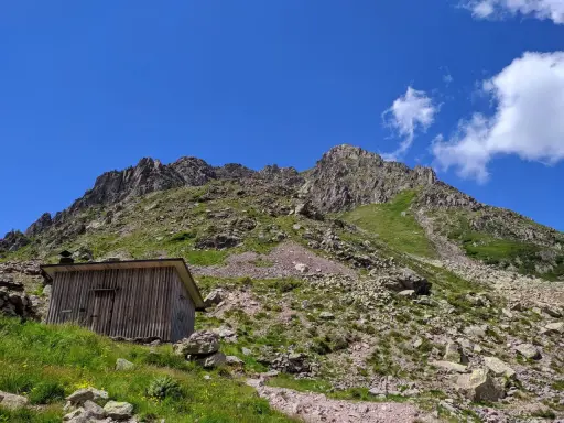 Bivacco Paolo e Nicola, un rifugio in legno ai piedi di montagne rocciose sotto un cielo azzurro.
