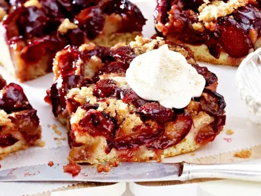 Saftiger Pflaumenkuchen mit Streuseln und einem Klecks Sahne. Ein Stück Kuchen liegt auf einem Messer, daneben eine Schale mit Sahne.