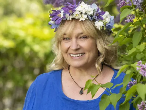 Sussie Eriksson med blomsterkrans i lila och vita blommor, en glad kvinna som påminner om hennes filmer och tv-program.