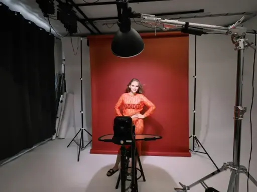 Modelka w pomarańczowej sukience pozuje na tle czerwonego tła. Wokół niej profesjonalne **światło studyjne** i sprzęt fotograficzny.