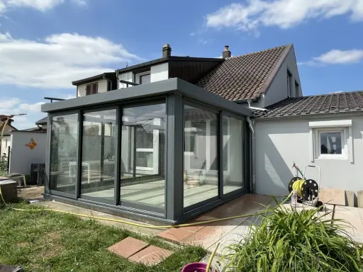 Une véranda moderne en verre et métal, prête pour l'assurance. Le soleil brille sur le jardin et la maison.