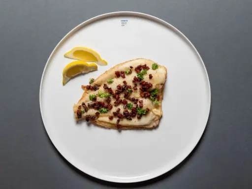 Scholle Finkenwerder Art: gebratenes Fischfilet mit Speckwürfeln und Petersilie, garniert mit Zitronenspalten auf einem weißen Teller.