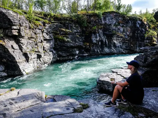 En kvinna sitter vid en forsande turkos flod i en klippig ravin. Vacker natur, kanske en vy över var ligger Abisko.