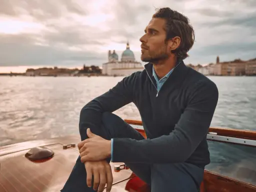 A man in mens dressy casual attire, a navy quarter-zip sweater over a blue collared shirt, sits on a boat with Venice in the background.
