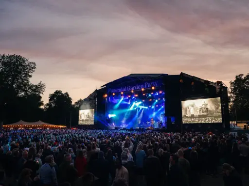 Stor publik vid Rosendal Garden Party. En scen med blå ljus och artister, omgiven av träd och en vacker kvällshimmel.