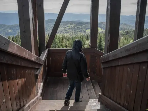 Osoba w czarnej kurtce schodzi po drewnianych schodach wieży widokowej. W oddali rozciąga się malowniczy krajobraz gór i lasów, który zapiera dech w piersiach.