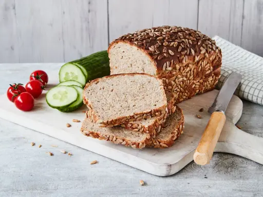 Frisch gebackenes Sonnenblumenbrot, zubereitet im Thermomix, liegt auf einem Holzbrett neben Tomaten und Gurkenscheiben.