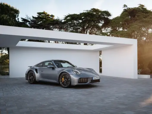 Silberner Porsche Turbo S, bereit für den 0-100 Sprint, parkt vor moderner Architektur im Sonnenuntergang.