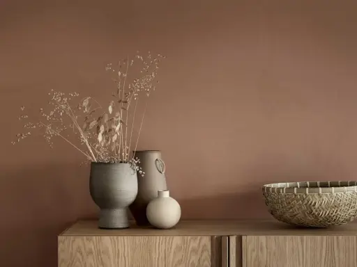 Tre vaser med torkade blommor och en flätad korg står på en träbyrå. En av vaserna är grå och har en rustik textur, en annan är beige med ett handtag och den minsta är rund och ljus.