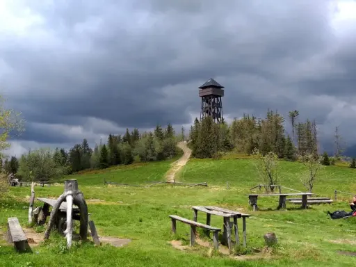 Drewniana wieża widokowa na Lubaniu. Trasa prowadzi przez zielone wzgórza pod burzowym niebem.
