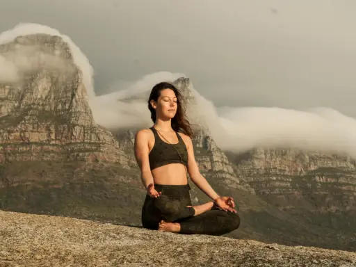 Kvinna i lotusställning på en klippa, med berg och moln i bakgrunden. Hon utövar buddhistisk meditation för inre lugn.