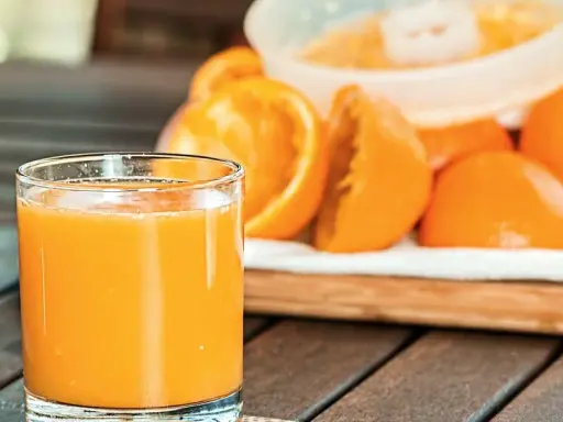 Szklanka świeżego soku pomarańczowego obok pokrojonych owoców, idealna na początek dnia lub jako element diety sokowej.