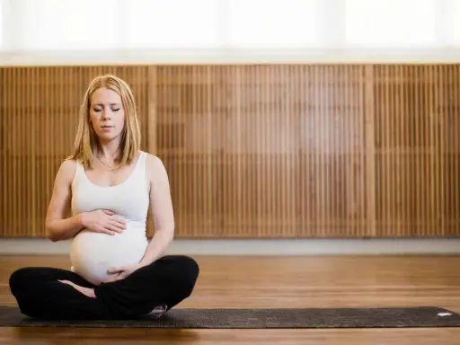 Gravid kvinna med slutna ögon sitter i lotusställning på en yogamatta. Hon håller sin mage, en lugn stund av gravid yoga.