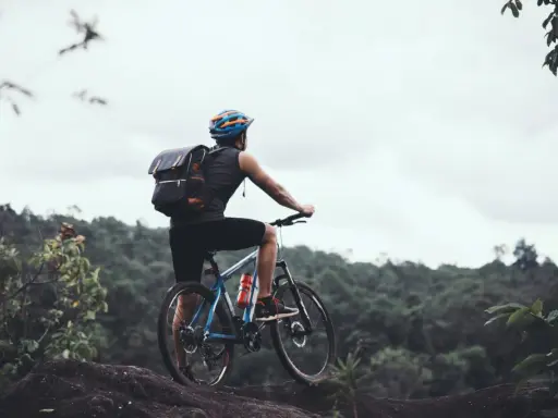 Rowerzysta z plecakiem na górskim szlaku, zastanawia się: gravel czy szosa? W tle zielone wzgórza i pochmurne niebo.