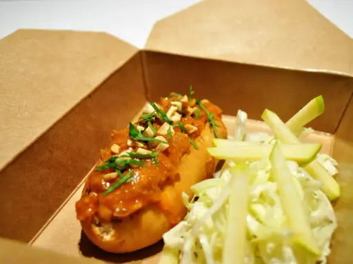 Un delicioso hot dog con salsa de cacahuete y cilantro, acompañado de ensalada de col y manzana, listo para tu delivery.