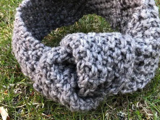 Grått, stickat pannband med en knut framtill, ligger på gräs. Perfekt för att sticka ett eget pannband.