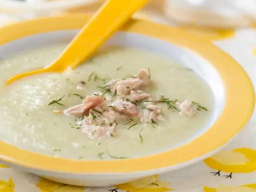Un cremoso puré de verduras con pollo desmenuzado y eneldo, servido en un plato amarillo con una cuchara.