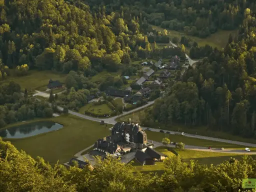 Widok z góry na osadę w lesie, przypominającą legendarną **Wilczą Górę**. Domy i hotel otoczone zielenią, z małym stawem.