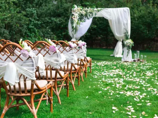 Eleganckie dekoracje na ślub cywilny w plenerze: rzędy krzeseł ozdobionych białymi wstęgami i kwiatami, łuk ślubny z tkaniną i zielenią, ścieżka usypana płatkami róż.