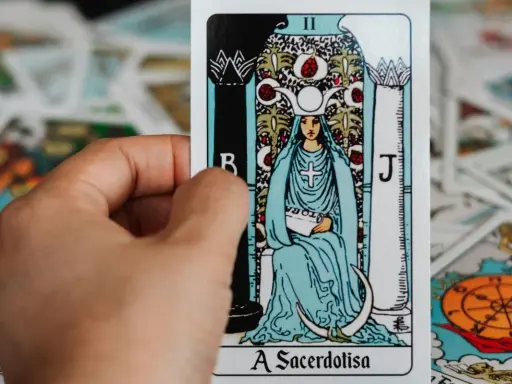 Dłoń trzyma kartę z tarot z 5 kart, przedstawiającą Wielką Kapłankę. W tle widać inne karty.