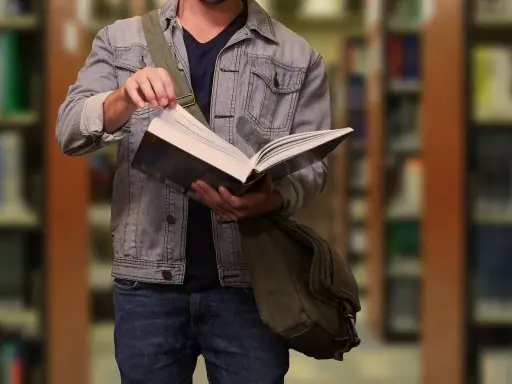 Student w dżinsowej kurtce przegląda książkę w bibliotece. W tle widać regały z książkami.