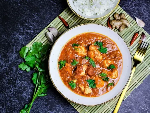 Ein Teller mit Hähnchen-Curry in cremiger **masala sauce**, garniert mit Koriander. Daneben Reis, Ingwer, Knoblauch und Chilis.
