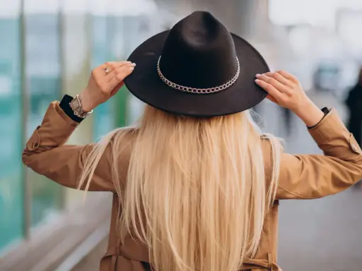 Blondynka w beżowym płaszczu i czarnym kapeluszu z ozdobnym łańcuszkiem. Pokazuje, jak nosić kapelusz, poprawiając go na głowie.