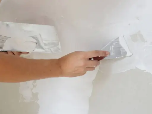 Persona aplicando masilla en una pared con dos espátulas. Aprende cómo masillar una pared para un acabado perfecto.