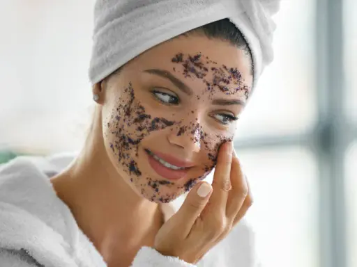 Une femme applique un gommage sur son visage, se demandant combien de gommage par semaine est idéal pour une peau éclatante. Elle porte une serviette sur la tête et un peignoir.