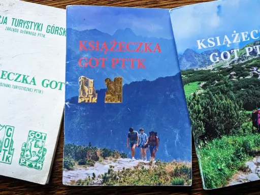Trzy książeczki GOT PTTK z odznakami. Jedna biała z zielonymi symbolami, dwie z górskimi krajobrazami.
