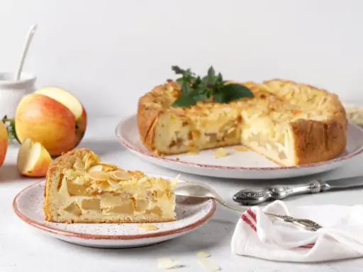 Un délicieux gâteau aux pommes, garni d'amandes effilées, est présenté avec une part déjà coupée. Des pommes fraîches sont disposées à côté.