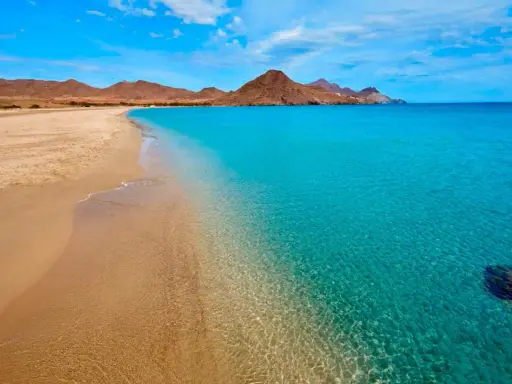 La **playa de los Genoveses** se extiende con arena dorada y aguas cristalinas de color turquesa, bajo un cielo azul con nubes blancas y montañas áridas al fondo.