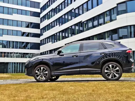 Czarny SUV MG HS, z nowoczesnym designem, zaparkowany przed biurowcem. Jego linia nadwozia sugeruje dynamiczne osiągi i niskie spalanie.