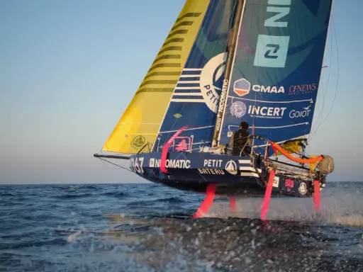 Segelboot mit pinken Foils rast über die Wellen. Ein Skipper steuert das Boot bei der Mini Transat Regatta.
