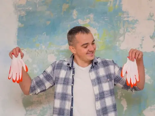 Homme souriant tenant des gants blancs à bouts orange devant un mur texturé. Il semble prêt à peindre, peut-être pour estimer le prix peinture m2 au black.
