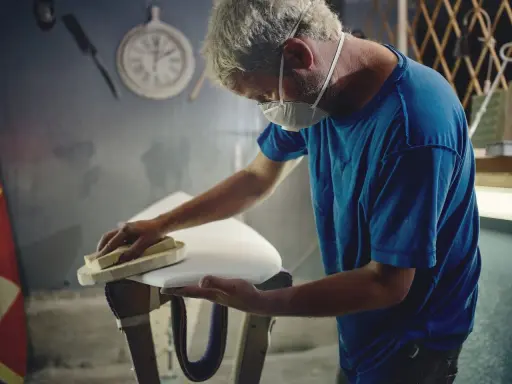 Artisan masqué ponçant une planche de surf blanche, étape cruciale avant de faire de l'époxy.
