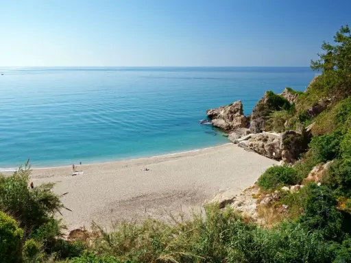 Playa de Burriana: Ein sonniger Strand mit türkisfarbenem Wasser, Felsen und üppiger Vegetation. Einige Menschen genießen die Ruhe.