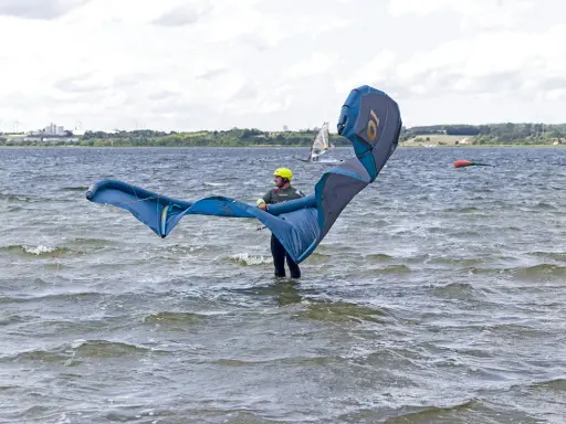 Osoba w żółtym kasku przygotowuje się do **kite surfingu**, trzymając duży, niebiesko-szary latawiec na wzburzonej wodzie.