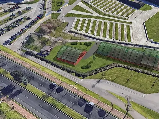 Vista aérea de un complejo deportivo con dos invernaderos verdes y una zona de aparcamiento. El **parque de las llamas** se extiende hasta la carretera.