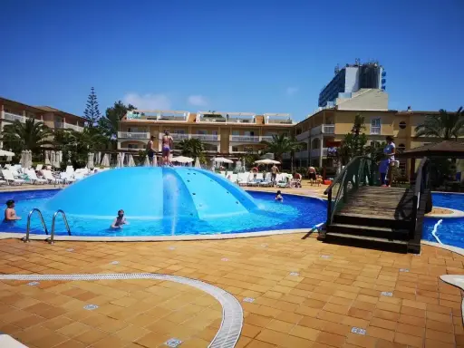 Sole e divertimento in piscina a Maiorca con bambini. Una grande struttura gonfiabile blu al centro, bagnanti e hotel sullo sfondo.