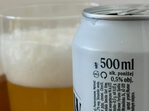 Piwo z puszki z etykietą "alk. poniżej 0,5% obj." obok szklanki z pianką. Sprawdzamy, ile promili ma to piwo.