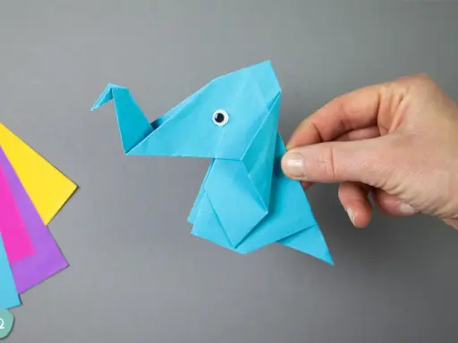 Ein blauer origami elefant mit einem kleinen Knopfauge wird von einer Hand gehalten. Daneben liegen bunte Papierbögen.