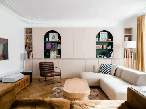 Un salon moderne avec un canapé beige, une chaise en cuir marron et des étagères murales roses. Quelle couleur pour une grande pièce à vivre ? Le vert émeraude des niches apporte une touche audacieuse.
