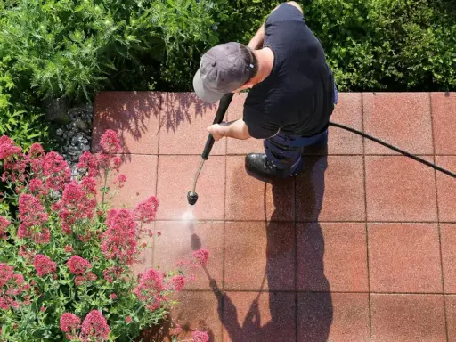 Mężczyzna w czapce z daszkiem czyści kostkę brukową myjką ciśnieniową, obok kwitnące różowe kwiaty i zielone krzewy.