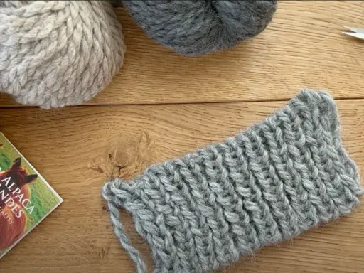 Ovillos de lana gris y beige sobre madera. Un trozo de tejido con puntos de lana muestra la textura.