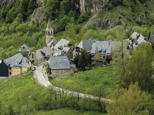 Pueblos del Valle de Arán, con casas de piedra y tejados grises, se asoman entre la vegetación verde y un camino serpenteante.